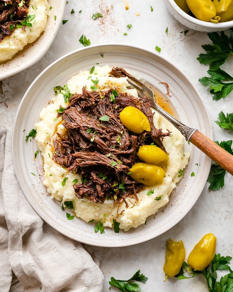 The dish is served on a white plate with a smooth, slightly ridged edge. The bottom layer is creamy mashed potatoes, spread evenly in a circular shape with a soft, fluffy texture in white color. On top of the mashed potatoes, there is a generous pile of shredded dark brown beef that looks tender and juicy, with some visible seasoning like black pepper. There are three yellow pickled peppers placed on the beef, adding a pop of bright color and a smooth, shiny surface. A sprinkling of fresh green chopped herbs is scattered over the meat and potatoes for a fresh look. A wooden-handled fork is positioned resting on the left side of the plate, slightly inserted into the potatoes. The background is a white marbled texture, with parts of another similar plate in the corner and scattered fresh parsley and yellow peppers nearby. photo taken with an iphone --ar 4:5 --v 7