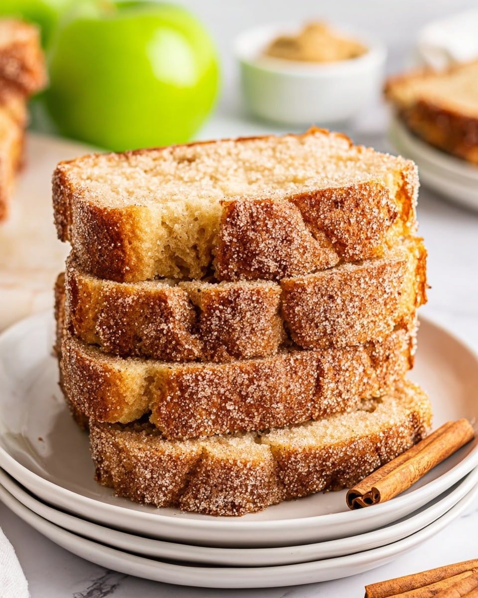 A stack of four thick slices of cinnamon sugar bread is placed on a white plate with two cinnamon sticks beside it on the right. Each slice shows a golden brown crust covered in a layer of sparkling cinnamon sugar, with a soft, slightly crumbly light brown interior. The plate is resting on another white plate, and the background features a white marbled texture with a green apple and a small white bowl of spread blurred in the distance. photo taken with an iphone --ar 4:5 --v 7