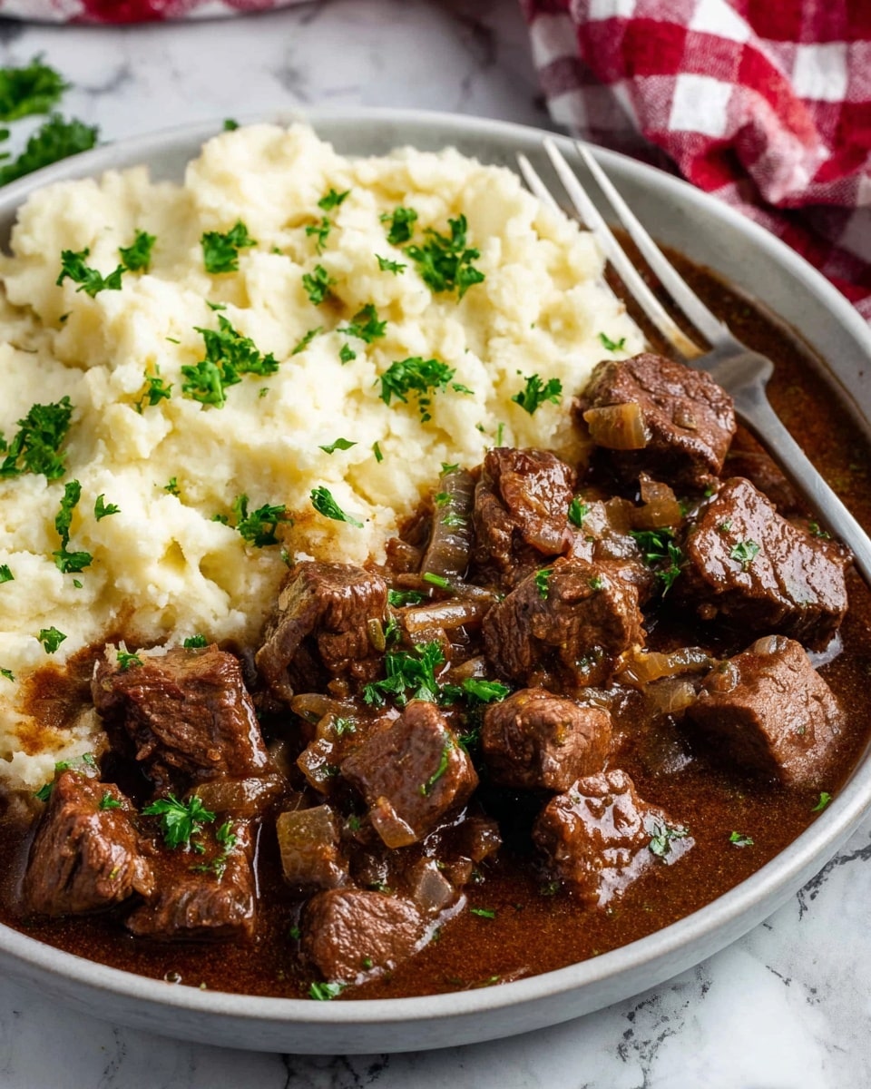 The dish is served on a white plate with two main layers: the left side has a thick, creamy white mashed potato layer with a few green parsley leaves scattered on top, and the right side is filled with chunks of brown beef stew in a rich, dark brown gravy with visible pieces of cooked onions and sprinkled green parsley. A fork rests on the plate's top edge over the stew, and the background shows a white marbled surface with a red and white checkered cloth slightly out of focus. Photo taken with an iphone --ar 4:5 --v 7
