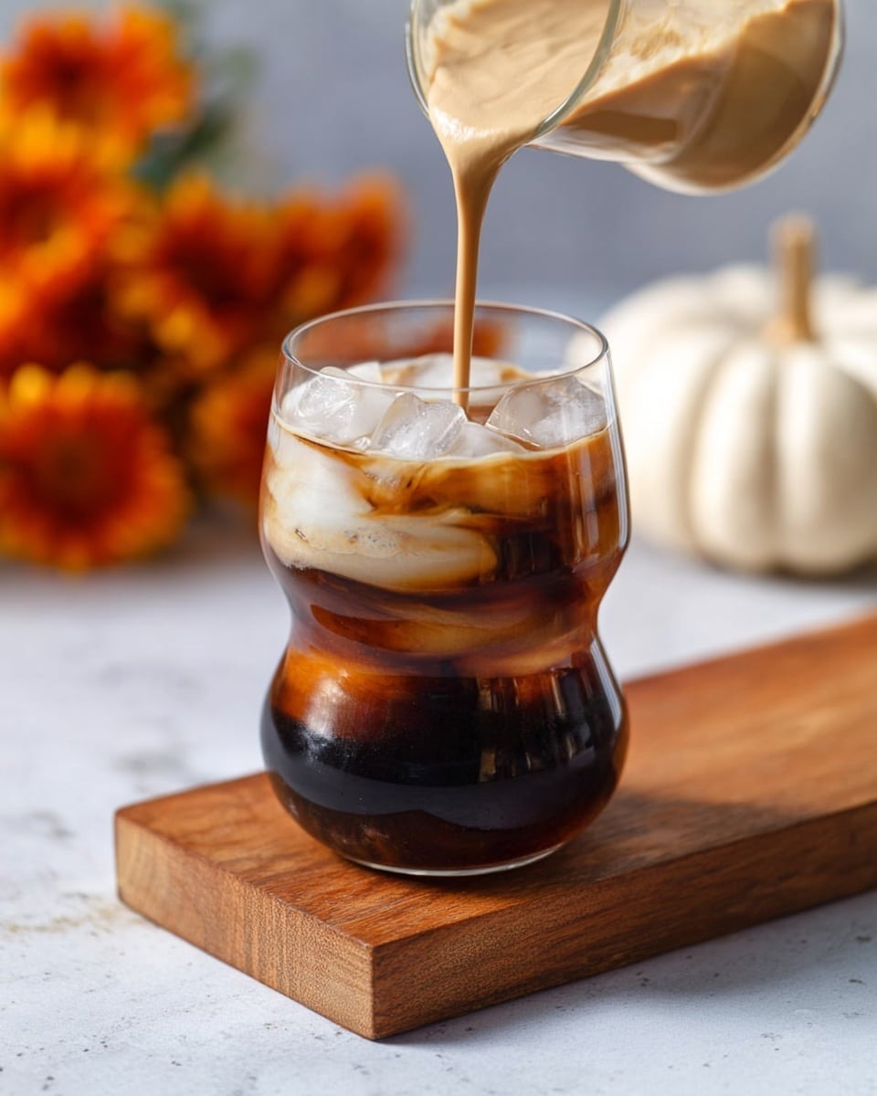 A clear glass with a unique curved shape holds a dark cold drink filled with ice cubes, showing three visible layers of ice and liquid that are deep brown and translucent. On top, a thick, creamy light brown layer is being poured into the glass, creating a smooth frothy texture that contrasts with the cold liquid below. The glass sits on a small wooden board, all placed on a white marbled texture surface, with blurry orange flowers and pumpkins in the soft background. photo taken with an iphone --ar 4:5 --v 7