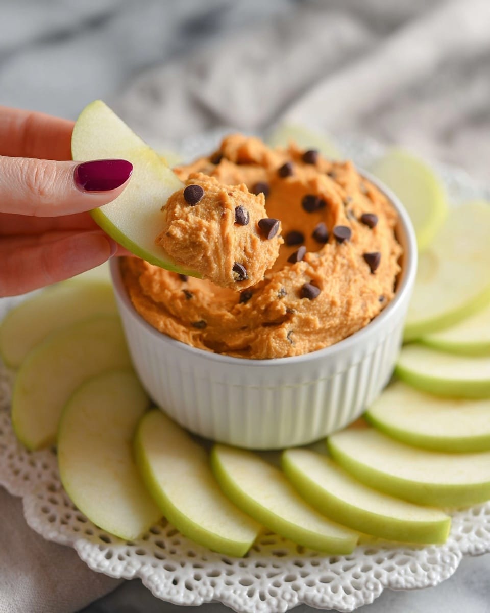 A small white ribbed bowl filled with thick, orange spread studded with small dark chocolate chips is centered on a white lace doily on a white marbled surface. Around the bowl, slices of green apple are arranged in a fan shape, showing their light green and pale yellow tones. In the foreground, a woman's hand with dark pink nail polish holds one apple slice dipped in the orange spread with chocolate chips clearly visible. The spread has a slightly rough texture with peaks and valleys from scooping. Photo taken with an iphone --ar 4:5 --v 7