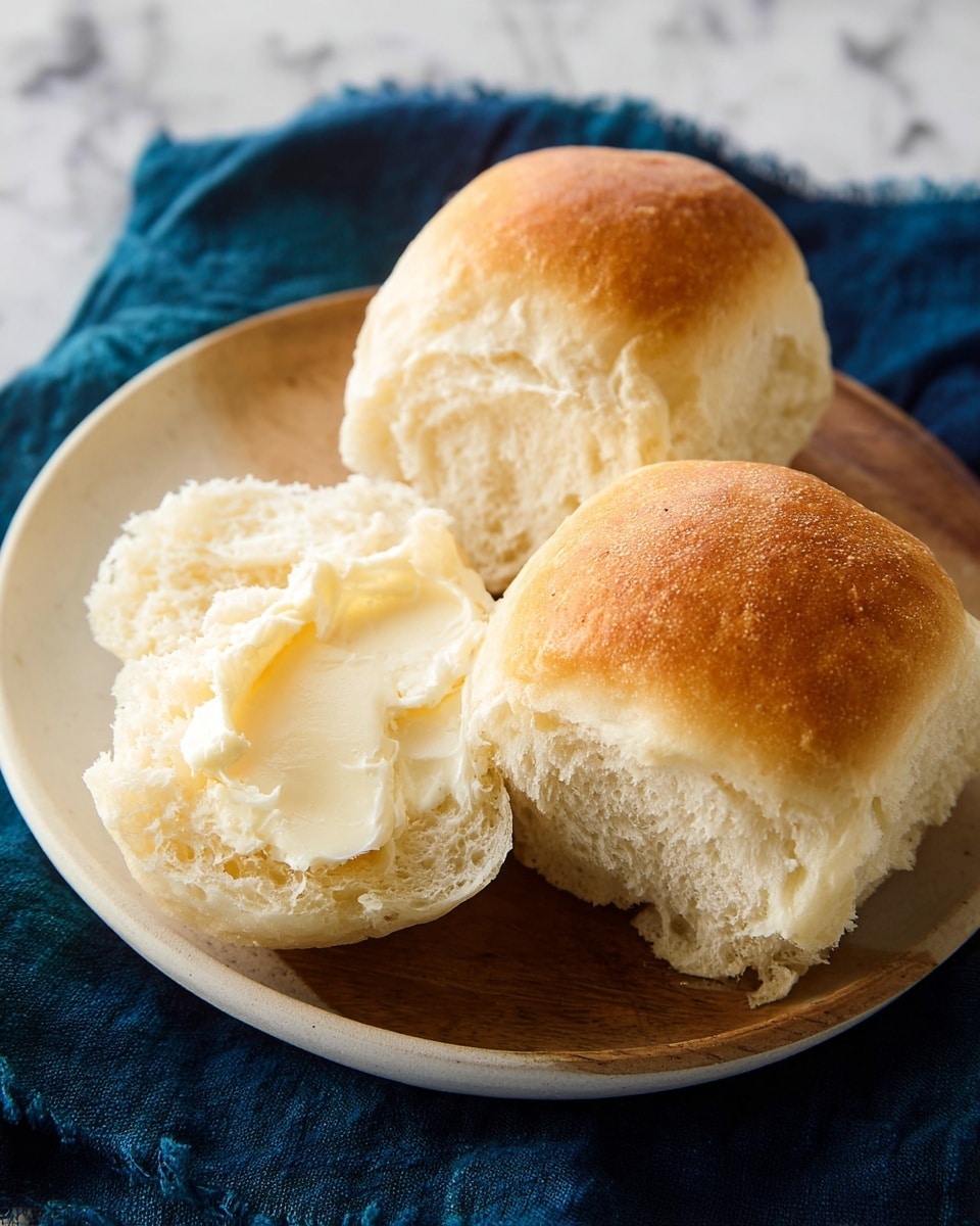 A white bowl filled with shiny golden brown dinner rolls stacked closely together, each roll showing a soft, fluffy light yellow inside and a smooth, glazed crust on top. The bowl is lined with a white cloth napkin with fringed edges, partly covering the rolls. In the background, a stack of white plates and a piece of cake are slightly visible, all set on a white marbled surface. photo taken with an iphone --ar 4:5 --v 7