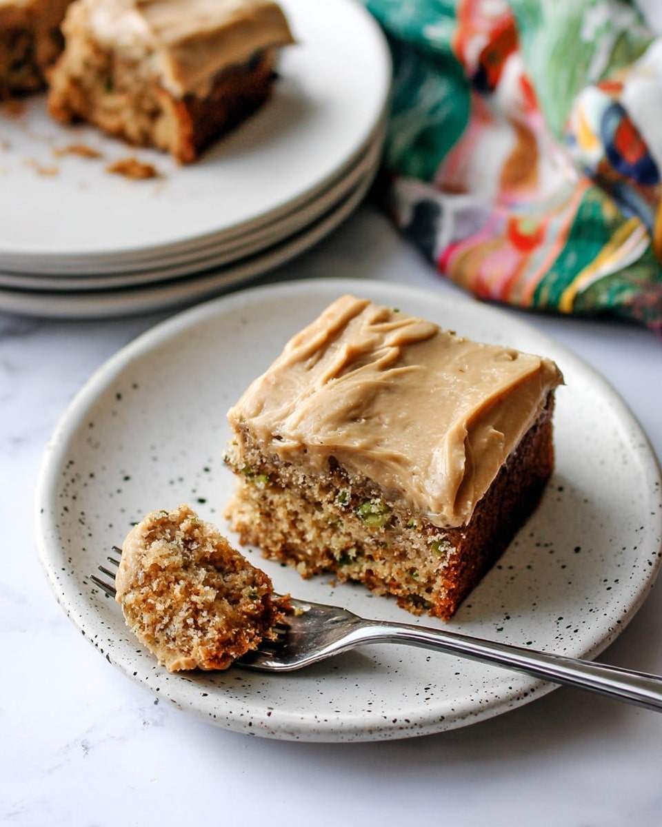 A tray of cake with a thick, smooth light brown frosting spread evenly on top, showing one piece missing from the corner. There are two slices of cake served on white speckled and plain white plates; the cake inside is light brown with visible small green bits, giving it a moist, crumbly texture. One slice on the white speckled plate has a fork with a bite of cake showing the inside texture clearly. The plates and tray are placed on a white marbled surface, with a folded floral cloth featuring soft purple, pink, and blue flowers next to some forks and knives. A few small green vegetable peelings lie scattered on the surface. photo taken with an iphone --ar 4:5 --v 7