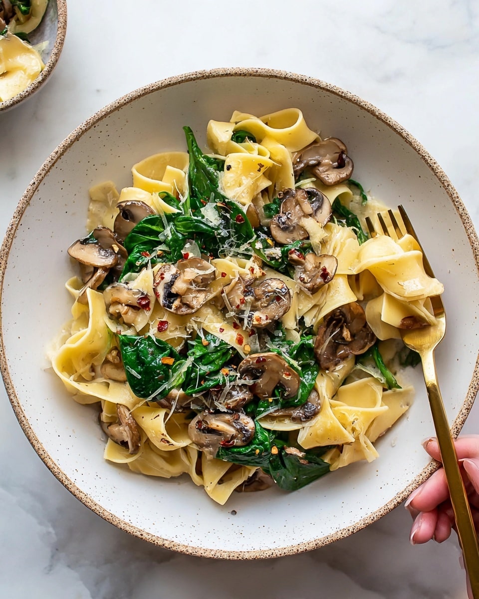 A bowl of wide, flat yellow pasta forms the base layer with a soft texture, topped with bright green spinach leaves that add freshness and color. Scattered on top are brown mushrooms, sliced and cooked, providing an earthy look and texture among the pasta and greens. Small bits of grated cheese and red chili flakes are sprinkled on the dish, adding white and red accents and a bit of texture contrast. A bronze fork is placed on the right side of the bowl, resting on some pasta, with a woman's hand holding it gently. The bowl is white with small brown speckles around the edges, all set on a white marbled surface. Photo taken with an iphone --ar 4:5 --v 7