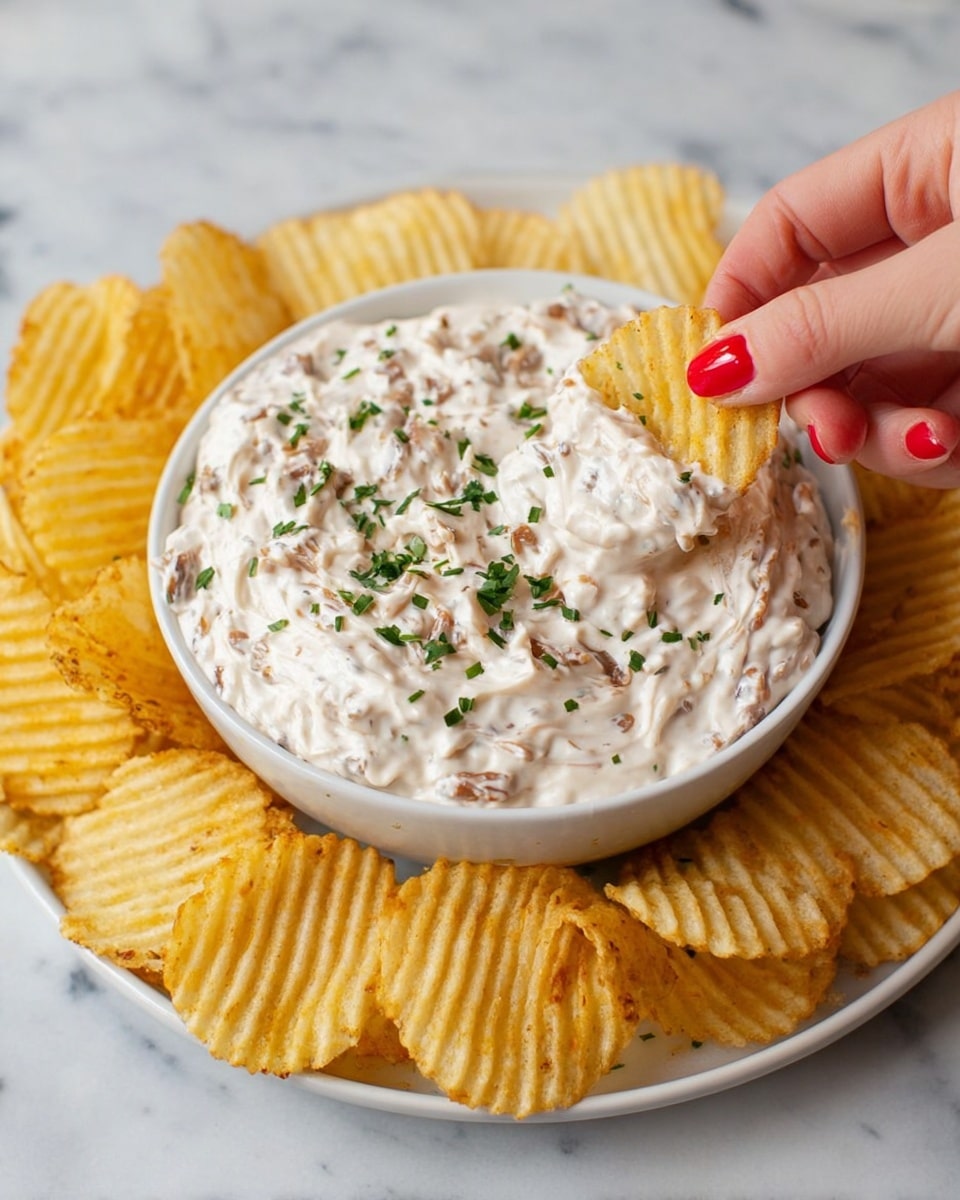 A white bowl filled with creamy, textured dip that has small pieces of browned bits mixed in, topped with chopped green herbs scattered on the surface; the bowl is placed in the center of a white plate, surrounded by a single layer of yellow ridged potato chips spread evenly around it on a white marbled surface. photo taken with an iphone --ar 4:5 --v 7