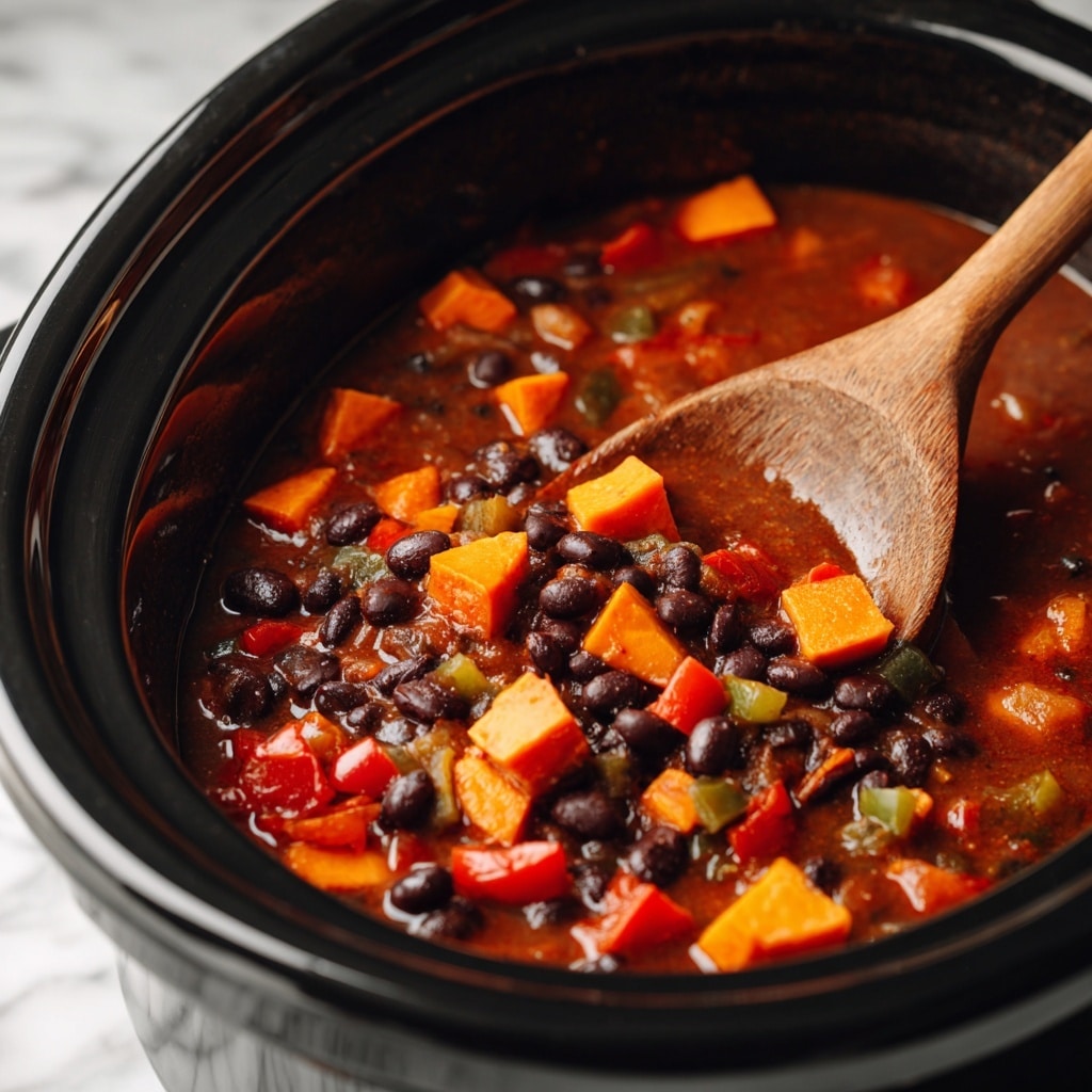 A close-up view inside a black slow cooker filled with a stew made of dark black beans, bright orange sweet potato chunks, diced red tomatoes, and small bits of green vegetables in a thick reddish-brown broth. A wooden spoon with a smooth grain rests inside the slow cooker, partially submerged in the stew, showing the chunky texture and colorful mix of ingredients against the black interior. The image background is a white marbled texture. Photo taken with an iphone --ar 4:5 --v 7