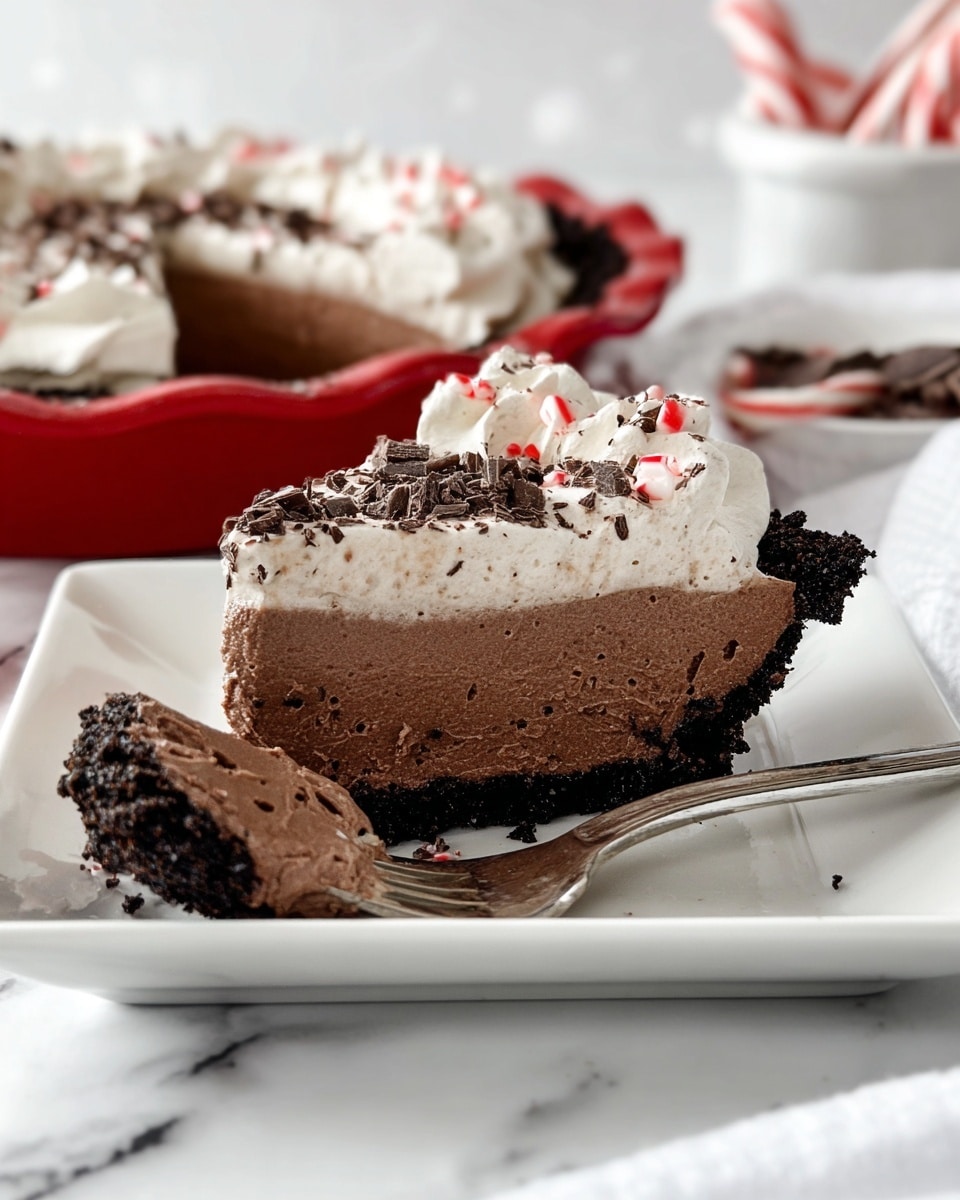 The image shows a slice of a chocolate cream pie with three visible layers on a white square plate over a white marbled surface. The bottom layer is a thick, dark, crumbly chocolate crust. The middle layer is dense, smooth, and dark chocolate mousse. The top layer is airy light chocolate whipped cream with small dark chocolate shavings and tiny red bits scattered over it. A small fork lies on the plate holding a bite-sized piece of the pie, clearly showing these three layers and textures. In the background, the rest of the pie sits in a red pie dish, with a white bowl and some candy canes partially blurred. Photo taken with an iphone --ar 4:5 --v 7