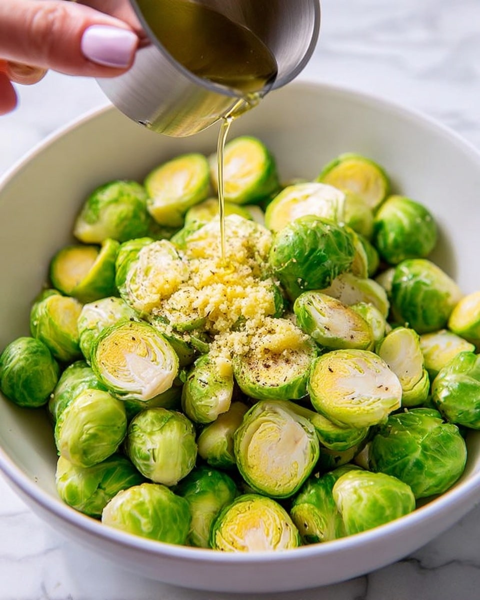 A close-up view of a white bowl filled with halved bright green Brussels sprouts, some showing a light yellow inside. On top of the sprouts, there is a small pile of light yellow crushed garlic and specks of black pepper sprinkled across. Above the bowl, a metal measuring cup is pouring a thin stream of golden olive oil onto the Brussels sprouts. A woman's hand with light pink nails holds the measuring cup. The bowl is set on a white marbled surface. photo taken with an iphone --ar 4:5 --v 7