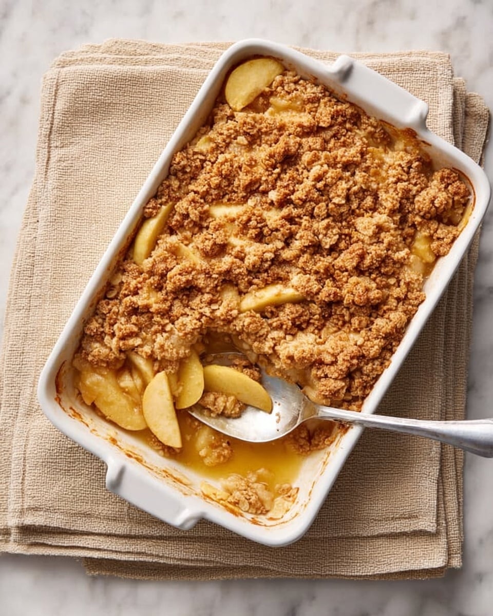 A white bowl filled with warm apple crumble topped with a single scoop of creamy vanilla ice cream. The bottom layer shows soft, golden apple slices coated with a light syrup, while the crumbly, textured top layer is golden brown with small clusters of baked flour and sugar. The ice cream on top is smooth and slightly melting into the crumble, adding a creamy contrast. The bowl is placed on a white marbled surface next to a shiny metal spoon and a beige striped cloth napkin, all lit by soft natural light. photo taken with an iphone --ar 4:5 --v 7