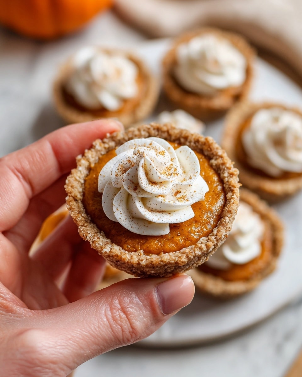 The image shows a small dessert with three layers. The bottom layer is a thick, rough-textured light brown crust shaped in a round, slightly raised edge form. The middle layer is a smooth, bright orange pumpkin filling sitting flat within the crust. The top layer is a small swirl of white whipped cream placed in the center of the pumpkin filling, sprinkled lightly with brown cinnamon powder. One of the desserts in the front is on a white, softly scalloped plate and has a bite taken out of it, showing the orange filling inside. In the background, more desserts are visible on a white plate, all placed on a wooden surface with a white marbled texture surrounding it. Photo taken with an iphone --ar 4:5 --v 7
