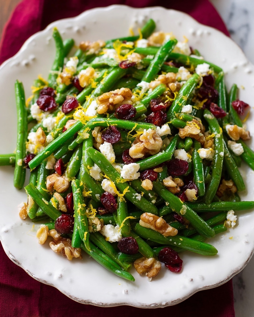 The image shows a white scalloped-edged plate filled with a green bean salad. The bottom layer is bright green beans cut into short pieces, with a smooth and slightly glossy texture. Scattered over the green beans are pieces of white crumbled cheese, rough and soft in texture. Mixed among these are small chunks of light brown walnuts, adding a rough and crunchy texture. Bright red dried cranberries are randomly placed throughout the salad, giving pops of deep red color. Thin yellow lemon zest strands are sprinkled on top, adding a fine, delicate texture. The plate sits on a white marbled surface with a dark red cloth slightly visible on the edges. Photo taken with an iphone --ar 4:5 --v 7