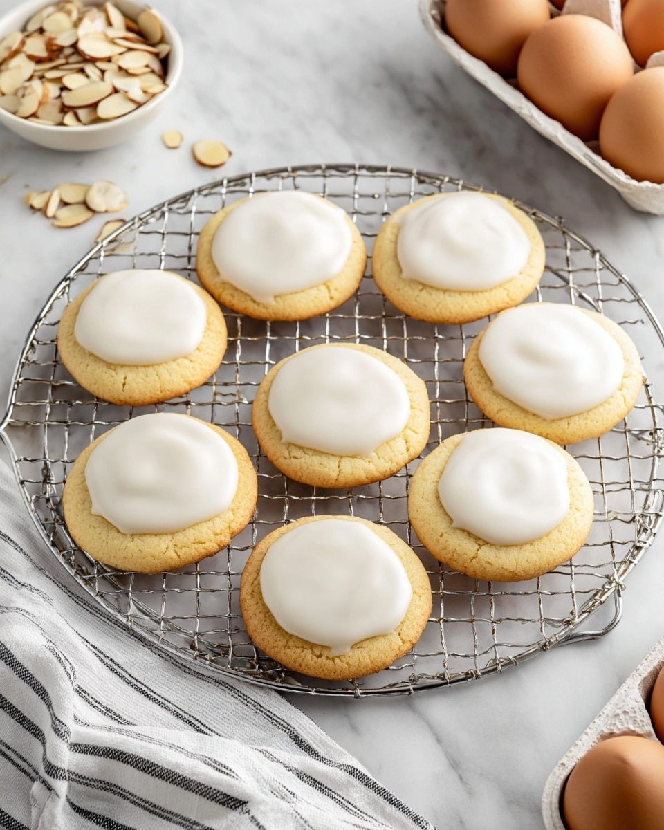 The image shows eight round cookies with a light golden color, each topped with a smooth, glossy white icing spread evenly in the center layer. They are placed on a round, metallic cooling rack with a grid pattern, positioned in the middle of a white marbled surface. Around the rack, there is a small white bowl filled with sliced almonds at the top right corner and a white egg carton holding four brown eggs at the bottom right corner. A striped white and black linen napkin is partially visible on the bottom left side, and one extra cookie sits directly on the white marbled surface near the bottom left of the rack. Photo taken with an iphone --ar 4:5 --v 7