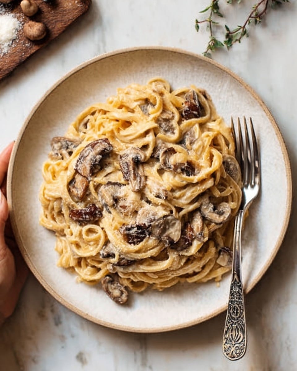 A close-up view of creamy pasta served in a white bowl filled with several layers of long fettuccine noodles mixed evenly with sliced mushrooms. The sauce is thick and light beige, coating the pasta smoothly, with small bits of herbs and black pepper sprinkled on top. The mushrooms are scattered throughout, showing a dark brown color and soft texture. The whole dish has a slight shine from the sauce, and some grated cheese is lightly dusted over the top. The bowl sits on a white marbled surface. Photo taken with an iphone --ar 4:5 --v 7