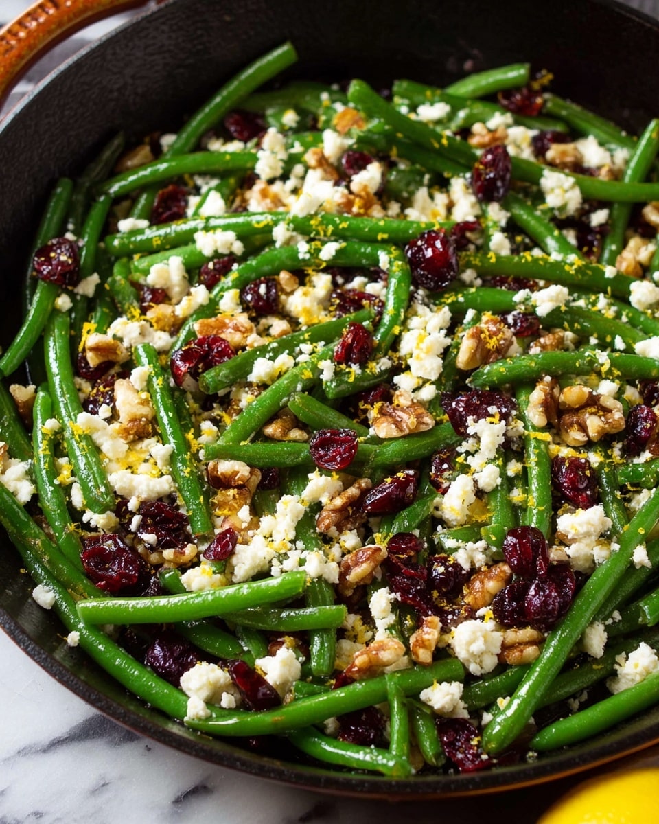 The image shows a close-up of a skillet filled with bright green cut green beans mixed with small white crumbled pieces of cheese, scattered red dried cranberries, and broken brown walnut pieces. The green beans form the base layer, fresh and vibrant with a slight shine, while the cheese is sprinkled unevenly on top, adding a soft, crumbly texture. The cranberries provide small pops of deep red color throughout, and the walnuts add a rough, crunchy texture with light and dark brown hues. Small bits of yellow lemon zest are also visible, adding a fine layer of color contrast on top. The skillet sits on a white marbled surface with a yellow lemon partially visible at the edge. photo taken with an iphone --ar 4:5 --v 7
