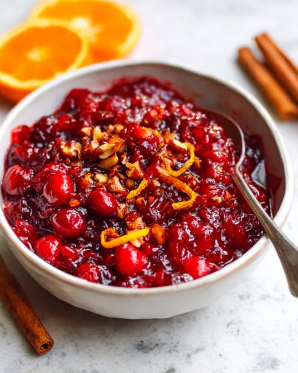 A white bowl filled with bright red cranberry sauce that has a thick, chunky texture. The sauce is mixed with whole cranberries, orange zest curls, and small pieces of nuts on top. A metal spoon rests inside the bowl, and the bowl sits on a white marbled surface with a sliced orange and a cinnamon stick nearby. The colors are warm and vibrant, with deep reds and bright orange details. Photo taken with an iphone --ar 4:5 --v 7
