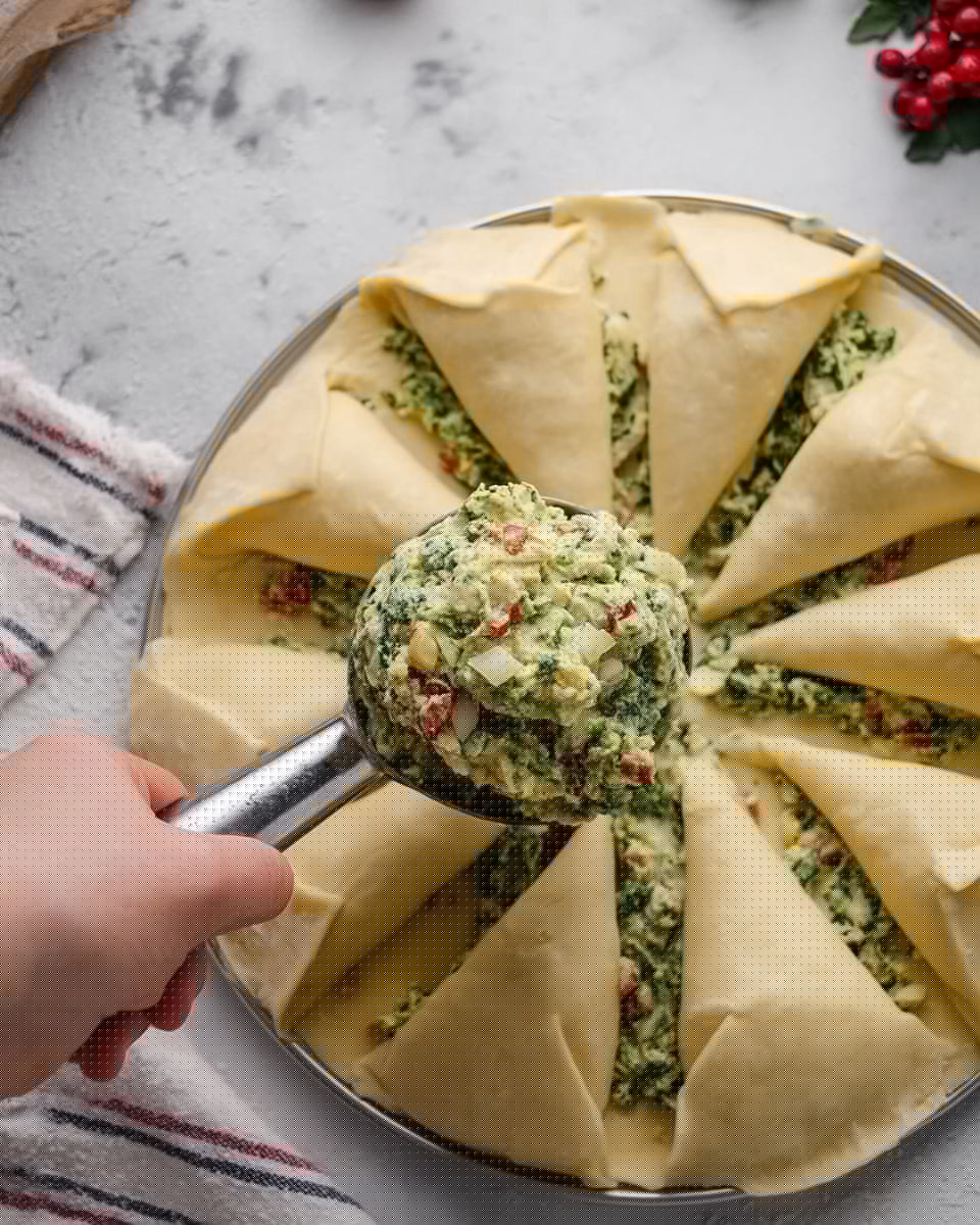 A ring-shaped bread wreath with a thick golden crust is topped with a layer of melted white cheese mixed with chopped green herbs. The wreath rests on brown parchment paper on a wooden cutting board, decorated with sprigs of fresh rosemary and red berries near the bottom. Around the board, there are green textured fabric, strings of red beads, a white bowl filled with dark red cranberries, and stacked white plates on a white marbled surface. Photo taken with an iphone --ar 4:5 --v 7