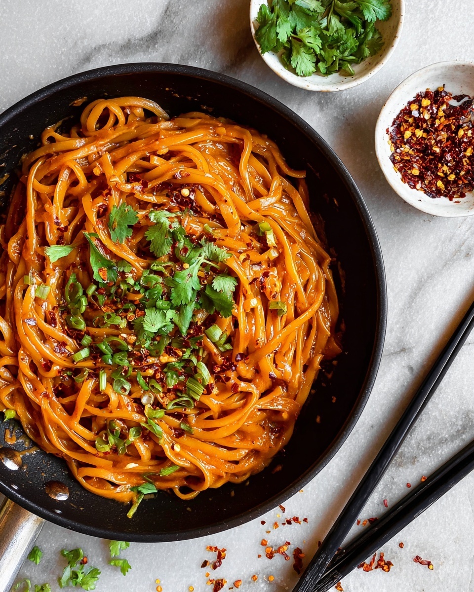 The image shows a black cast iron skillet filled with creamy reddish-orange noodles coated in sauce. The noodles are thick and flat, layered evenly across the skillet. Fresh green cilantro leaves are sprinkled on top, adding a bright contrast. Around the skillet, there are two small white bowls, one with chopped green herbs and the other with red chili flakes. Black chopsticks rest on the white marbled surface next to the skillet. Small bits of chili flakes and cilantro are scattered lightly on the surface. photo taken with an iphone --ar 4:5 --v 7