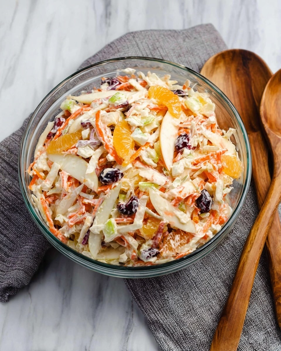 A clear glass bowl filled with a colorful salad made of several layers of thinly sliced and shredded ingredients, including white apple slices, orange segments, shredded carrots, small dark berries, and light green pieces of celery or cucumber, all mixed evenly together. The salad has a creamy texture, visible as a light coating over the ingredients. The bowl sits on a white marbled surface next to a gray cloth and two wooden salad utensils with smooth, polished handles. photo taken with an iphone --ar 4:5 --v 7