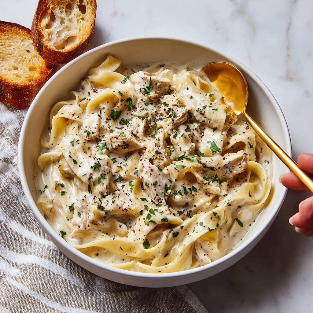 A large white pot is filled with wide, flat pasta noodles coated in a creamy white sauce with visible black pepper specks spread evenly throughout. Small chunks of light-colored chicken are scattered among the noodles, blending smoothly with the sauce. A dark wooden spoon partially dips into the sauce on the right side, lifting some noodles and sauce. The background shows a white marbled surface with a textured beige and white cloth partially visible underneath the pot. photo taken with an iphone --ar 4:5 --v 7