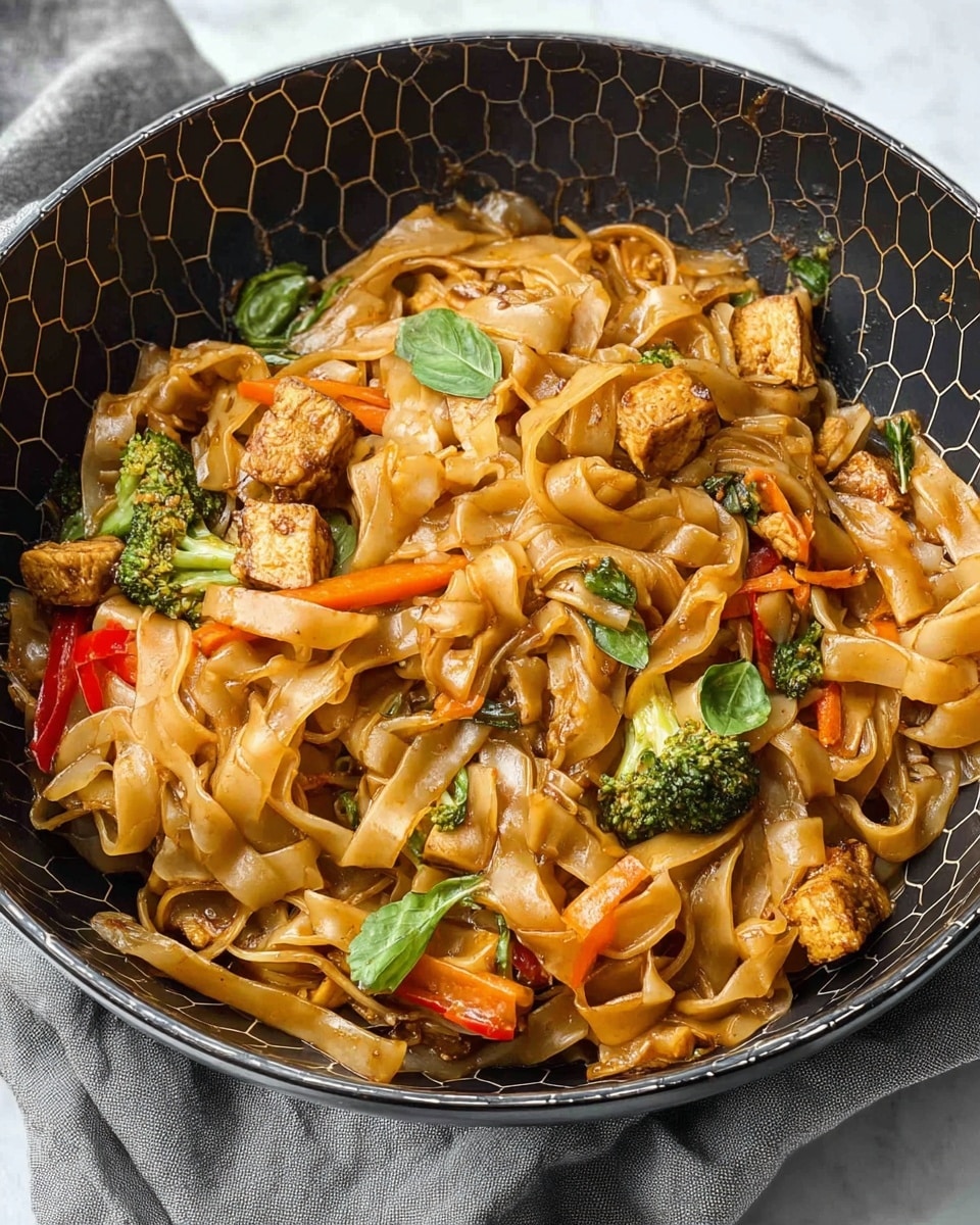 A large white bowl filled with wide, flat brown noodles mixed with pieces of golden-brown cooked chicken, bright green broccoli florets, slices of red bell pepper, and thin orange carrot strips. The noodles are coated in a glossy, dark soy-based sauce, giving them a shiny texture. The vegetables add pops of color, with the broccoli's green and peppers' red standing out against the brown noodles. The dish looks hot and fresh with steam rising slightly. It sits on a white marbled surface with some green leaves beside the bowl and a gray cloth partially visible. photo taken with an iphone --ar 4:5 --v 7