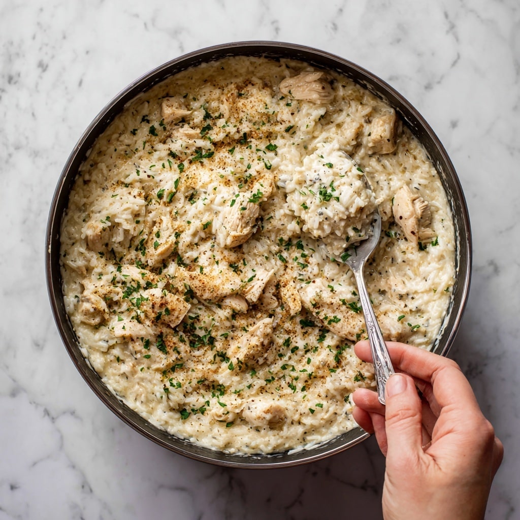 A white bowl filled with creamy chicken and rice, where the dish has a soft, thick texture with chunks of tender chicken mixed in. The top is sprinkled with finely chopped green herbs. A woman's hand holds a silver spoon scooping a generous spoonful from the bowl, showing the mix of smooth rice and chicken pieces. The bowl sits on a soft textured cloth over a white marbled surface. photo taken with an iphone --ar 4:5 --v 7