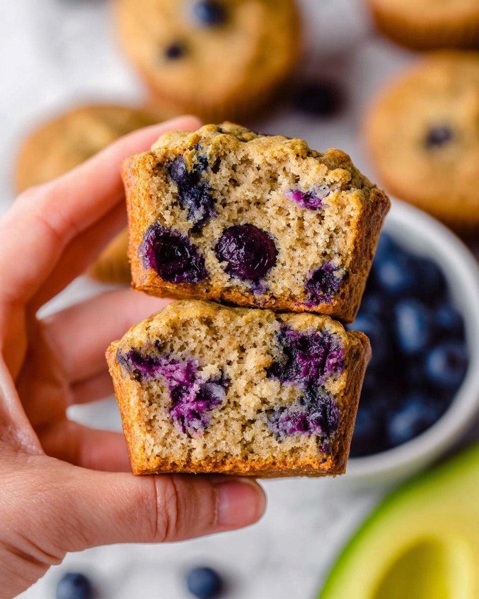 The image shows a cooling rack filled with golden-brown blueberry muffins that have a rough, slightly cracked top texture, each dotted with whole, dark blue blueberries. Among the muffins, there are loose blueberries scattered around and a halved avocado with smooth light green flesh and a brown seed on the right side. A white bowl filled with fresh blueberries is placed in the top left corner. The background is a white marbled texture. photo taken with an iphone --ar 4:5 --v 7
