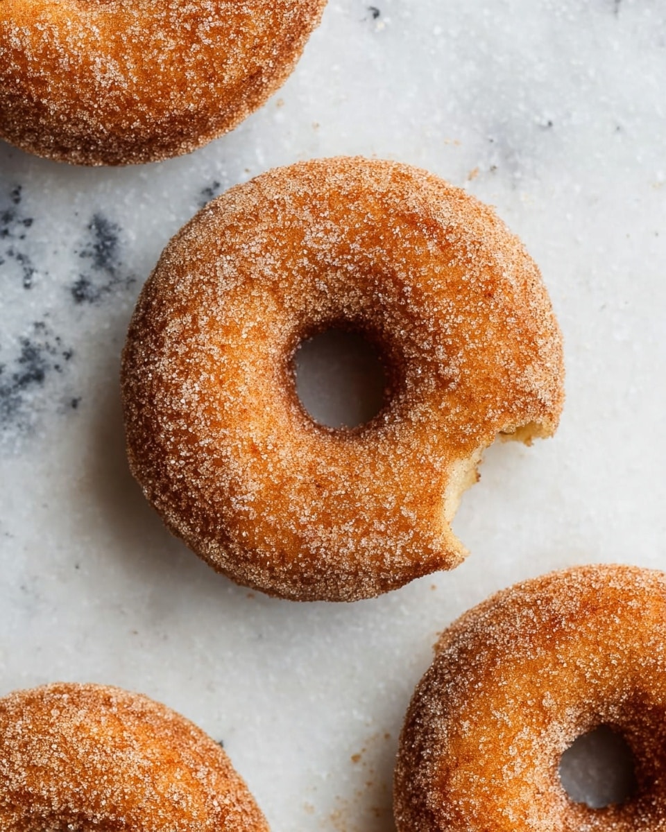 The image shows three sugar-coated donuts placed on a white marbled surface. Each donut has a golden-brown color with a rough texture covered evenly with granulated sugar. One donut is in the center with a small bite taken out from its outer edge, showing the soft, light inside contrasting with the crispy sugary coating. The other two donuts are positioned in the top left and top right corners, partly cropped by the frame, maintaining the same golden texture and sugar coating. photo taken with an iphone --ar 4:5 --v 7