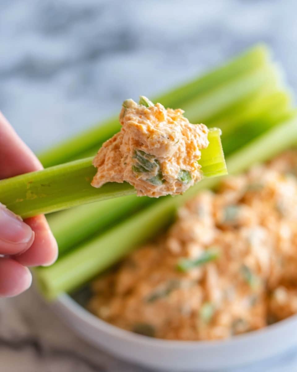A white bowl filled with a creamy, textured orange chicken salad mixed with small green celery pieces on top, behind it several fresh, bright green celery sticks are laid out. The bowl sits on a soft gray cloth over a white marbled surface, showing a close-up view that highlights the creamy and chunky layers of the chicken salad with smooth celery sticks in the background. photo taken with an iphone --ar 4:5 --v 7