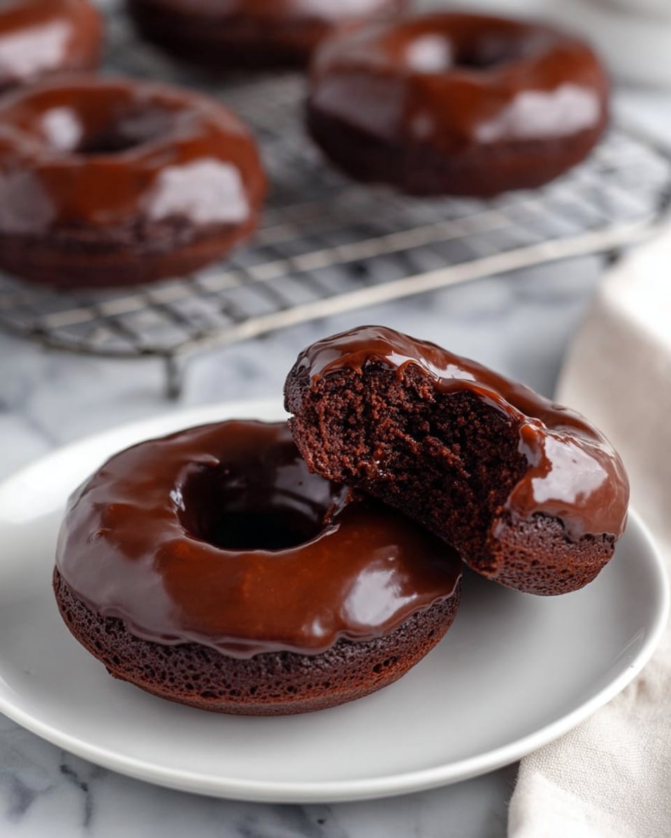 Four chocolate-glazed donuts sit on a golden wire cooling rack over a white marbled surface. Each donut has a thick, shiny dark chocolate glaze covering it smoothly; two donuts have extra fine chocolate shavings sprinkled on one side while the other two have thin chocolate drizzle lines on top. Part of a white towel with brown spots is seen in the lower left corner, and a bowl filled with rich chocolate sauce is placed in the upper left corner. Photo taken with an iphone --ar 4:5 --v 7