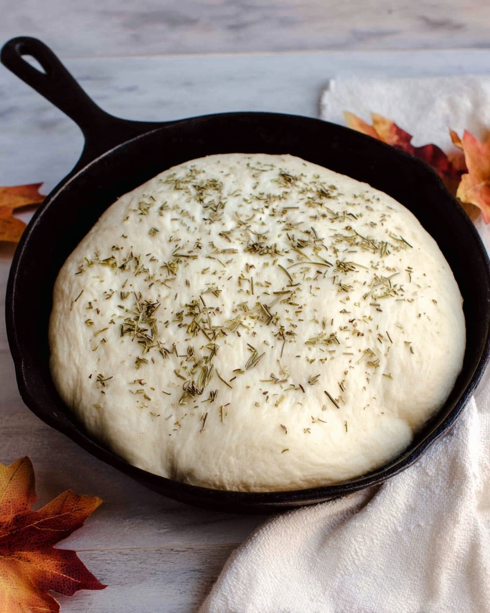 A round loaf of bread with a golden-brown crust sits in a black cast iron pan. The bread has eight thick slices scored on the top, showing a fluffy white inside beneath the shiny surface. It is sprinkled with small pieces of chopped garlic and thin green rosemary leaves scattered evenly over the top. Three green rosemary sprigs lie in the center for decoration. The pan rests on a gray cloth on a white marbled surface. Photo taken with an iphone --ar 4:5 --v 7