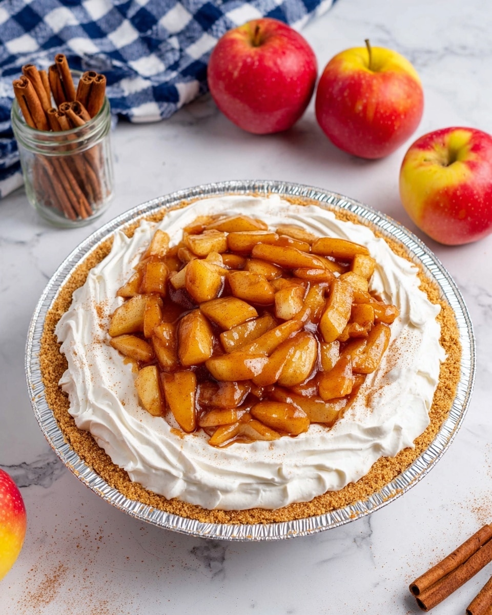 The image shows a pie with three clear layers: a light brown crumb crust forming the base, a thick white whipped cream layer in the middle spread evenly with soft swirls, and a shiny caramelized apple topping arranged in chunky slices in the center, glistening with a sticky glaze and sprinkled lightly with cinnamon. The pie is in a round foil pan and sits on a white marbled surface. Around it are three red and yellow apples, a glass jar with cinnamon sticks, a couple of loose cinnamon sticks, and part of a blue and white checkered cloth. Photo taken with an iphone --ar 4:5 --v 7