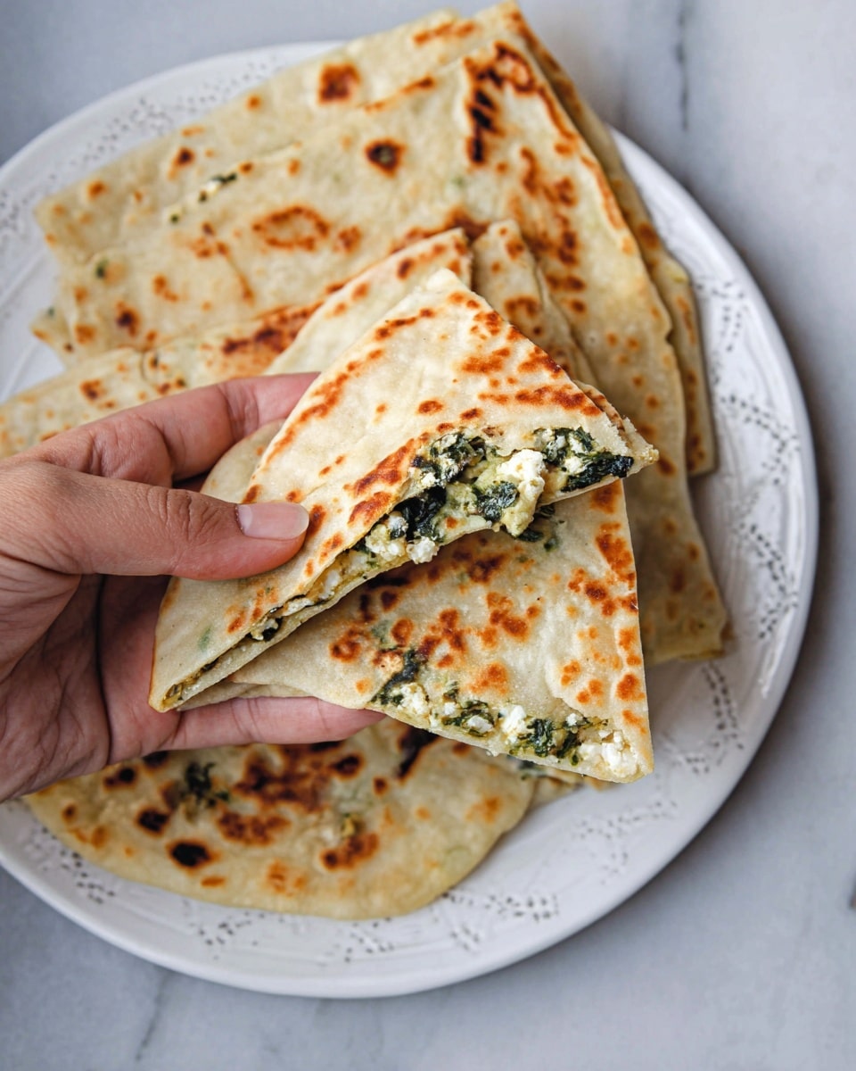 The image shows a stack of four folded flatbreads placed on a round silver plate. The flatbreads are golden brown with darker toasted spots on the surface. One flatbread is folded to reveal a filling of white cheese mixed with dark green leafy vegetables. A piece of this flatbread is cut out and lays on the white marbled surface next to the plate, showing the cheese and vegetable filling inside. In the background, there is a brown clay cup filled with a white frothy drink. The setting is simple with a wooden wall behind and a white marbled surface below. Photo taken with an iphone --ar 4:5 --v 7