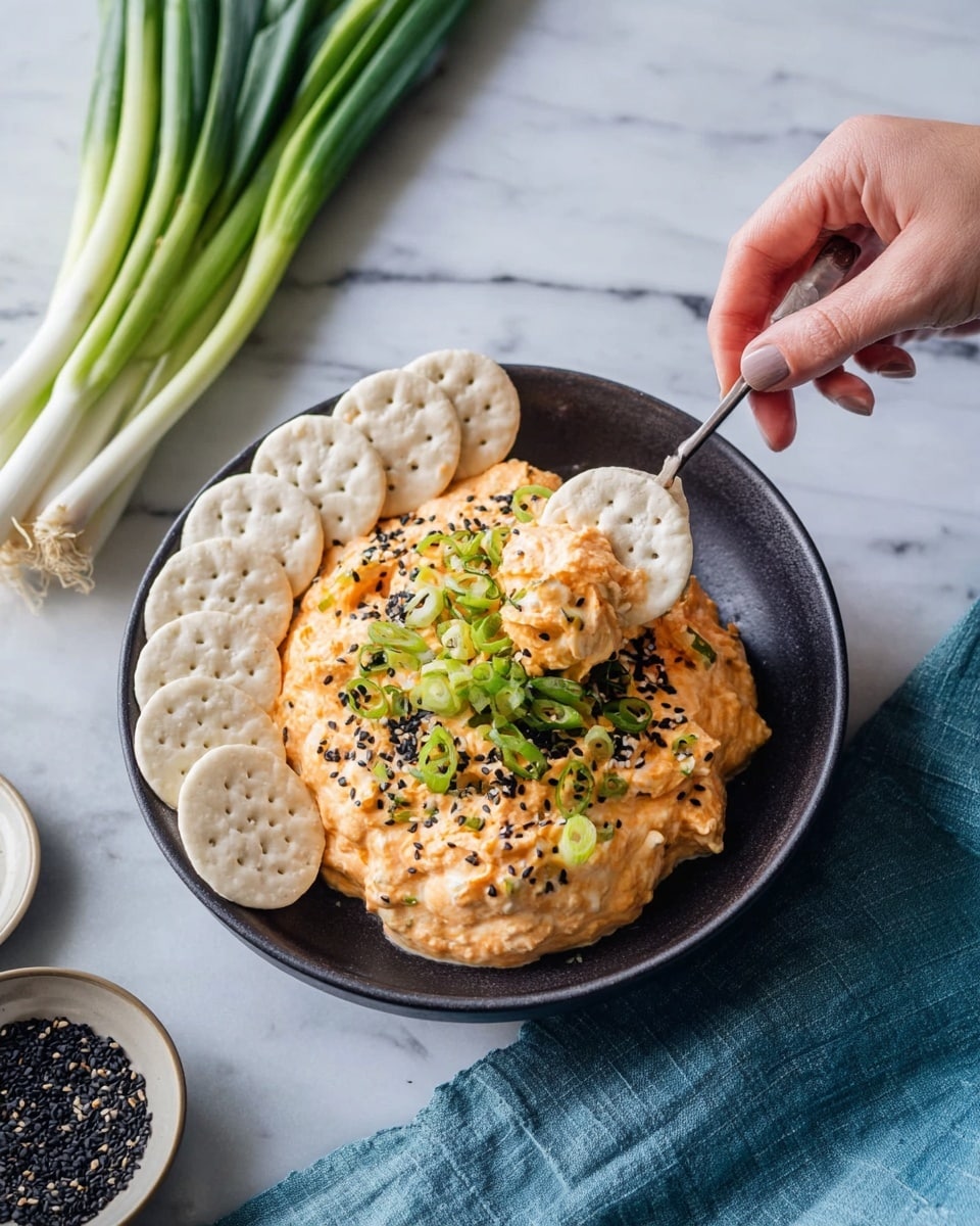 A black plate on a white marbled surface holds a creamy orange spread with visible chunks of white and pink, topped with green chopped scallions and small black sesame seeds. On one side of the plate, there is a neat row of round, white crackers with a slightly rough texture. A silver spoon rests partially in the spread, showing its soft and creamy texture. In the background, some fresh green scallions and a blue cloth are slightly blurred. photo taken with an iphone --ar 4:5 --v 7