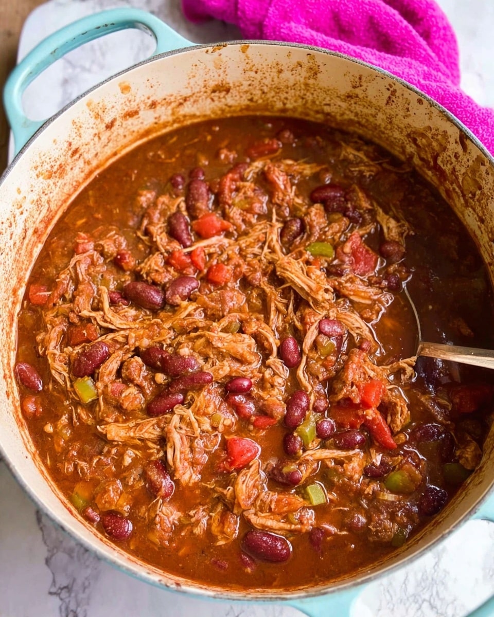 A close-up view inside a light blue white pot filled with a thick, chunky stew consisting of shredded meat, red kidney beans, diced tomatoes, and small pieces of green and red vegetables, all mixed in a rich brownish-red sauce; the sides of the pot show some sauce stains, and a metal spoon rests partially submerged on the right side of the stew, with a bright pink towel in the background on a white marbled surface. photo taken with an iphone --ar 4:5 --v 7