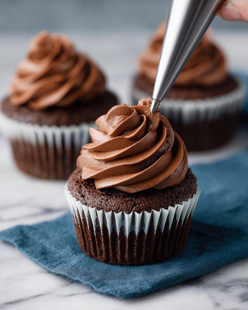 The image shows a close-up of a single chocolate cupcake on a white marbled surface with a dark blue cloth underneath. The cupcake has a dark brown base with a white paper liner and is being topped with smooth, creamy chocolate frosting swirled in a thick, even spiral. Two more similarly frosted chocolate cupcakes are blurred in the background, placed on the same dark blue cloth. A woman's hand is piping the frosting from a clear piping bag with a metal tip, adding texture and shape to the soft frosting. The overall look is rich and smooth with a focus on the swirl details of the frosting. photo taken with an iphone --ar 4:5 --v 7
