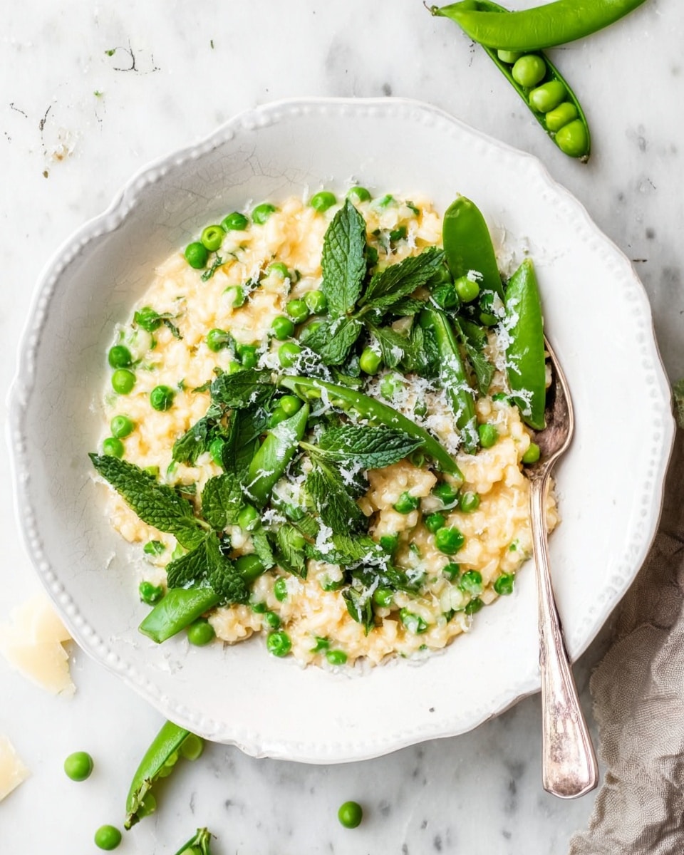 A white scalloped bowl filled with creamy yellow risotto as the base layer, scattered with bright green peas throughout. On top, there is a fresh layer of halved snap peas and whole peas, adding crisp texture and deeper green color. Fresh mint leaves are mixed on the top, along with a fine sprinkling of grated white cheese, giving a light textured snow-like effect. A silver spoon rests on the right side inside the bowl. The bowl sits on a white marbled surface with some loose peas, snap pea pods, and white grated cheese around it. Photo taken with an iphone --ar 4:5 --v 7
