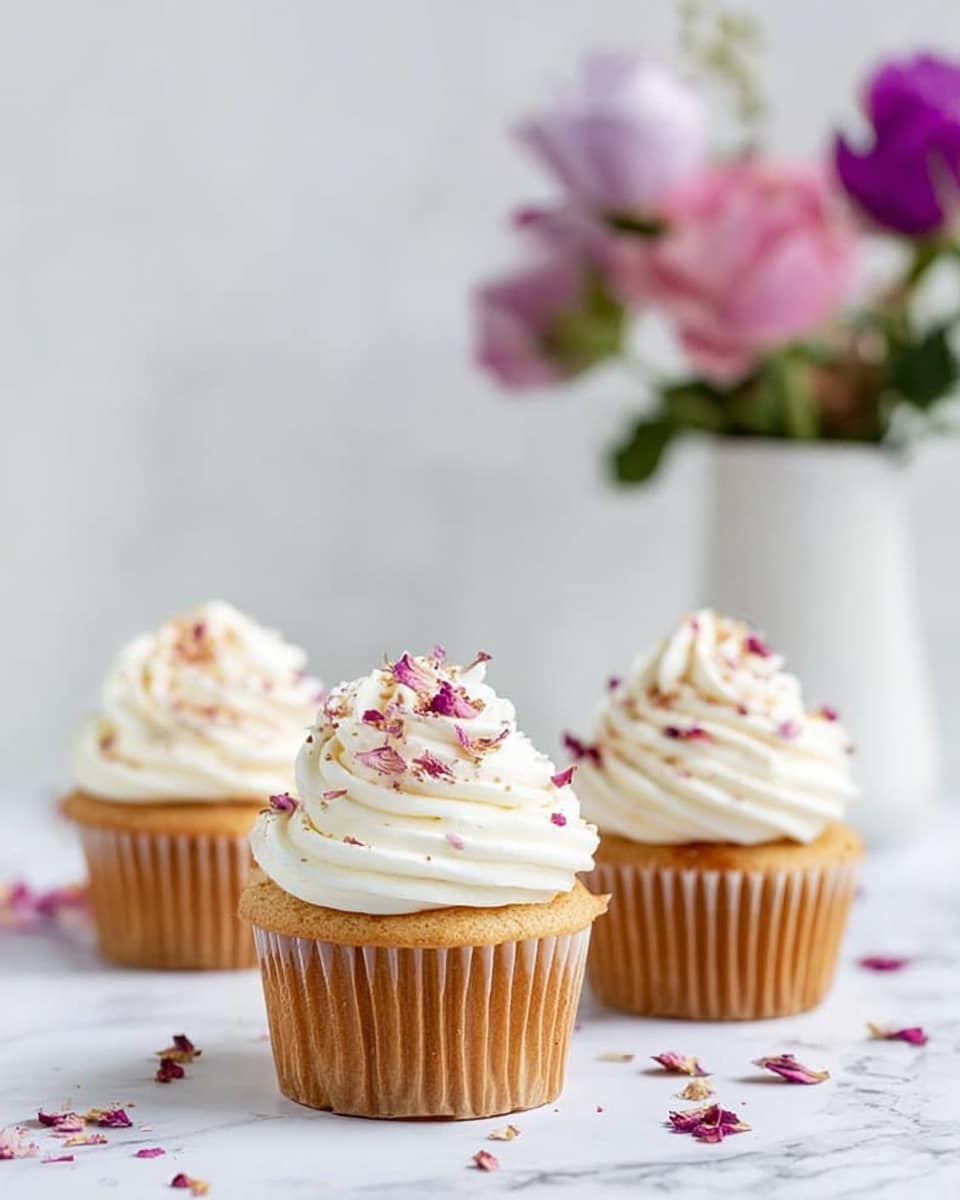 The image shows three cupcakes with light brown bases and tall swirls of white frosting on top. Each frosting swirl is decorated with small, colorful flower petal bits scattered across the surface. The cupcakes are placed on a white marbled texture, with a small white bowl containing more flower petals in the background. Soft pink flowers lie slightly out of focus behind the cupcakes, adding a gentle touch to the scene. Photo taken with an iphone --ar 4:5 --v 7