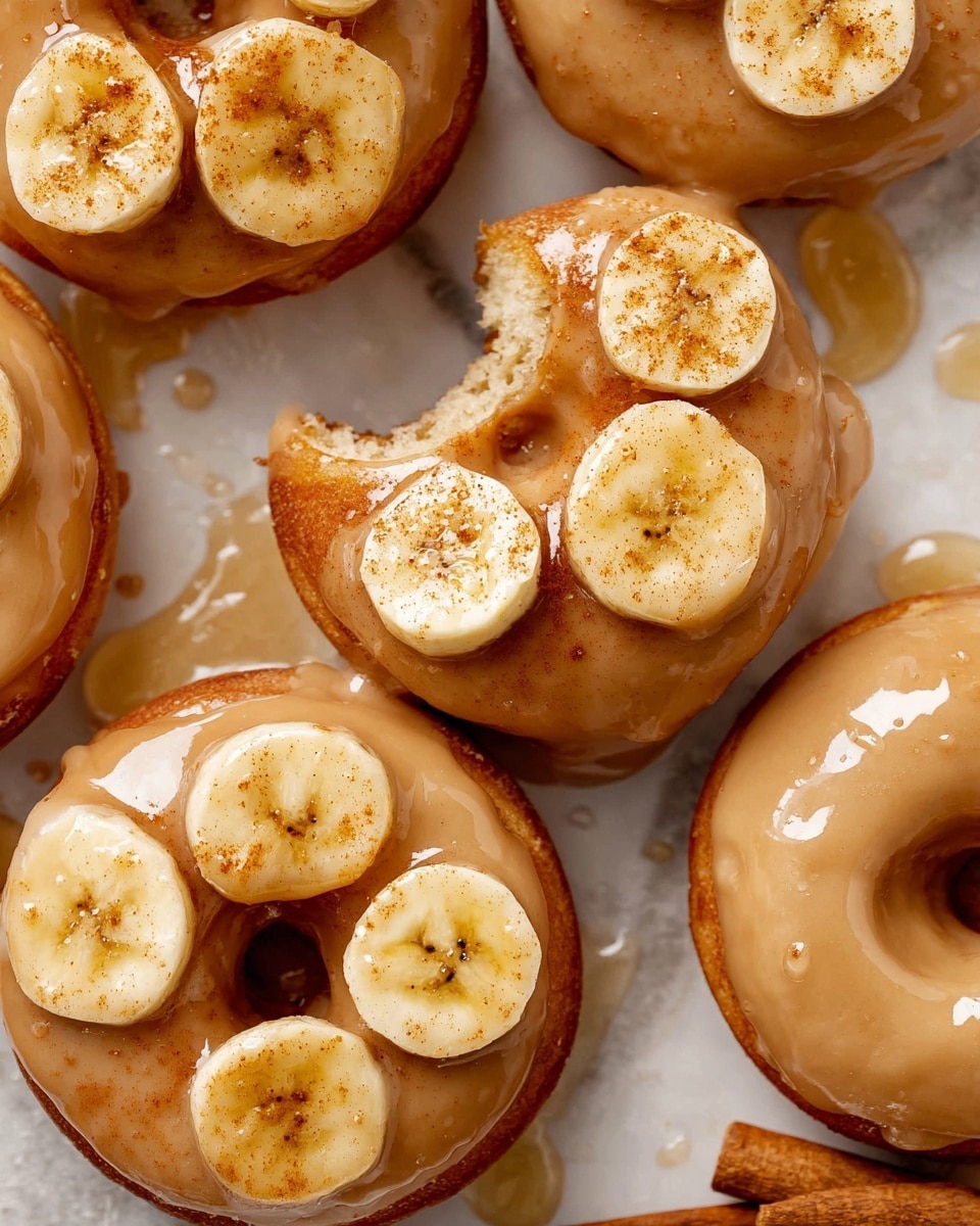A close-up of small round donuts stacked on a piece of parchment paper on a white marbled surface, each donut showing two layers with a light brown dough base and a thick, glossy caramel glaze on top. The top layer shines with a smooth caramel coating and has thin, yellow banana slices glazed and sprinkled lightly with cinnamon. One donut is in focus with a deep bite taken from it, showing a soft, airy inside with a light tan color. The background is softly blurred, showing more donuts. photo taken with an iphone --ar 4:5 --v 7
