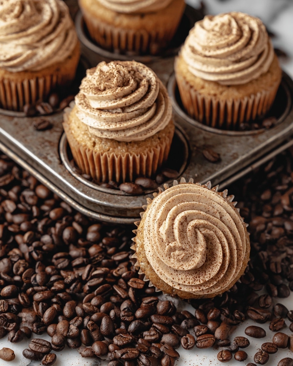 A close-up image of five cupcakes placed in a vintage metal cupcake tray, surrounded by many dark brown coffee beans scattered over a white marbled texture. Each cupcake has a single layer of golden-brown cake with a ridged paper liner and is topped with a thick swirl of light brown frosting, dusted lightly with coffee powder. One cupcake is pulled out a bit, resting at an angle on the edge of the tray, showing the detailed texture of the cake and frosting. The coffee beans fill the tray holes and spill around the tray, adding contrast with their shiny, smooth surface. photo taken with an iphone --ar 4:5 --v 7
