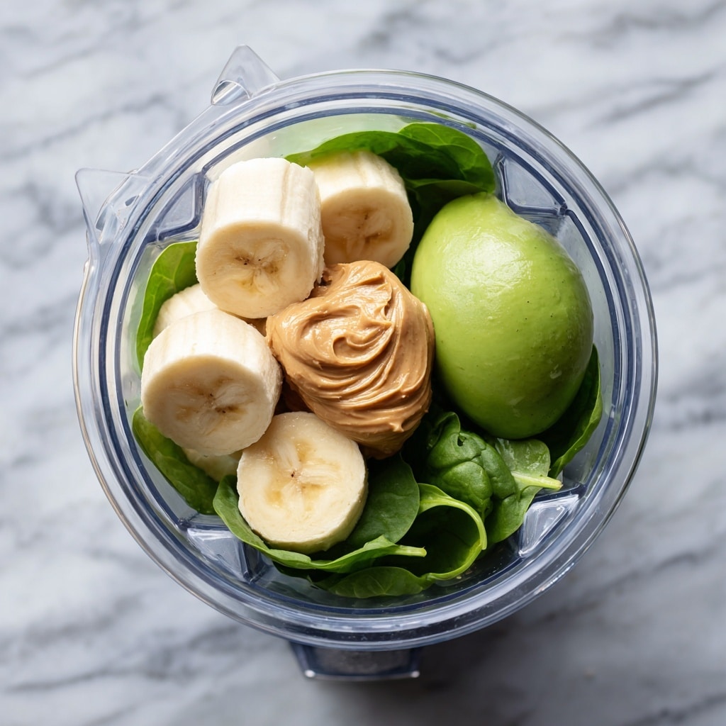 A clear glass filled with a thick, smooth green smoothie with tiny bubbles on the surface, garnished with a thin banana slice on the rim, and a white straw inside. The glass has a ribbed texture and sits on a white rectangular marble slab on a white marbled texture background. Around the glass are parts of fresh ingredients including a halved avocado, half a lemon, a spinach leaf, and a blurred second glass of the same smoothie in the background. Photo taken with an iphone --ar 4:5 --v 7
