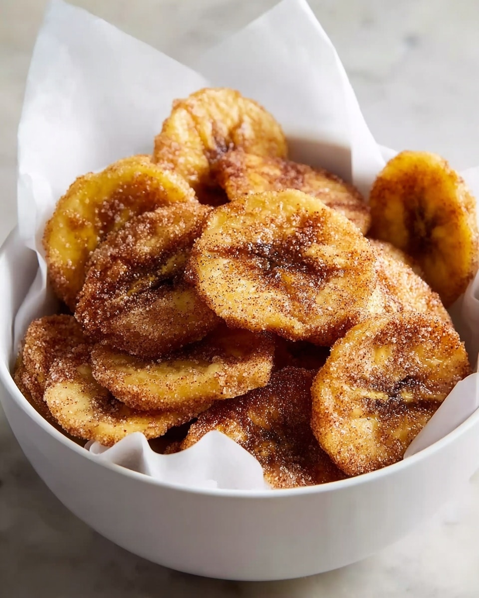 A white bowl lined with white paper holds several thin, round slices of fried plantains. Each slice has a golden-yellow base with a slightly crispy, brown cinnamon-sugar coating sprinkled all over, giving the pieces a textured look. The slices are layered loosely, filling the bowl nearly to the top, showing both the flat and curved edges, and creating a warm and crunchy appearance. The bowl sits on a white marbled texture surface. photo taken with an iphone --ar 4:5 --v 7