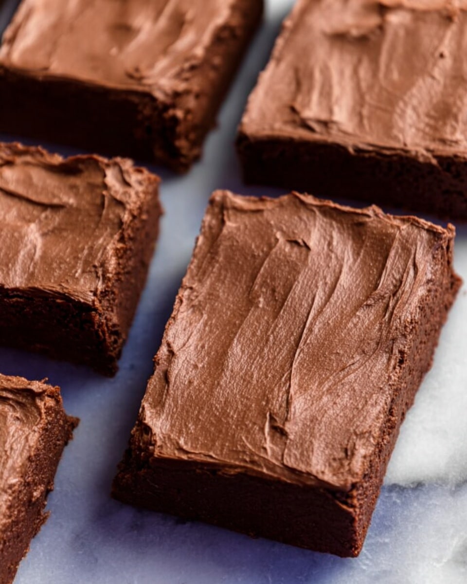 A stack of four thick chocolate brownies with a smooth, shiny chocolate frosting on top and between each layer, showing a rich, dense texture. The brownies sit on a piece of parchment paper that rests on a white marbled surface. In the background, more brownies are scattered flat, with the top layer also covered in the same glossy chocolate frosting. Photo taken with an iphone --ar 4:5 --v 7
