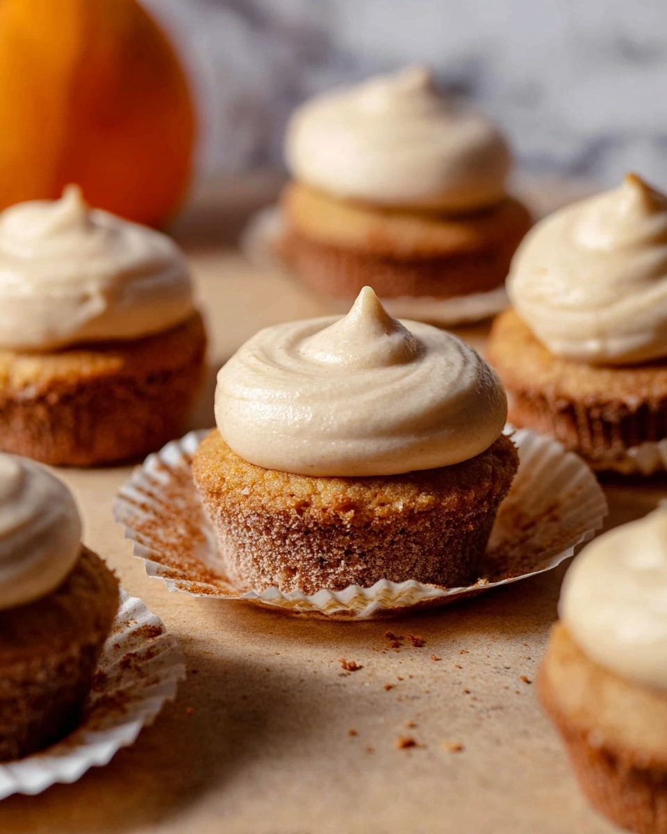 The image shows several small cupcakes, each with two visible layers: a bottom layer of moist, brownish-orange cake with a slightly rough texture, and a top layer of smooth, light beige frosting that has a small peak in the middle. Some cupcakes sit in white paper liners that are partially peeled away, revealing the textured cake beneath. The cupcakes are arranged closely together on a light brown surface with a few scattered crumbs. The background features a blurred round orange object, hinting at a pumpkin, and the whole scene is set against a white marbled texture. Photo taken with an iphone --ar 4:5 --v 7