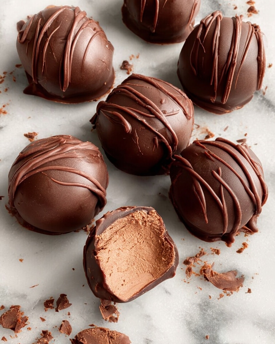 A white bowl filled with round chocolate truffles, each coated in smooth, shiny milk chocolate with some having a drizzle pattern on top for texture. One truffle is cut in half and placed on top, showing a creamy light brown filling inside with a firm outer chocolate shell. The bowl sits on a white marbled surface with a soft pink towel in the background. photo taken with an iphone --ar 4:5 --v 7