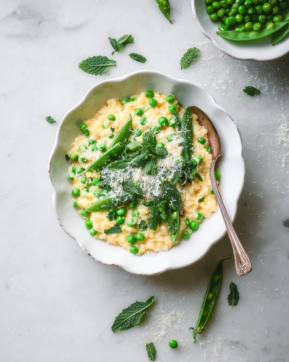 A white scalloped bowl filled with creamy, light yellow risotto mixed with bright green peas scattered throughout. On top, there is a generous layer of fresh snap peas, some whole and some sliced open to show the small peas inside, along with fresh green mint leaves adding a leafy texture. The dish is sprinkled with fine white shreds of cheese, creating a subtle contrast on the creamy risotto. A silver fork rests on the right side inside the bowl. The bowl sits on a white marbled surface with some loose peas, snap pea pods, and cheese shreds around it. photo taken with an iphone --ar 4:5 --v 7