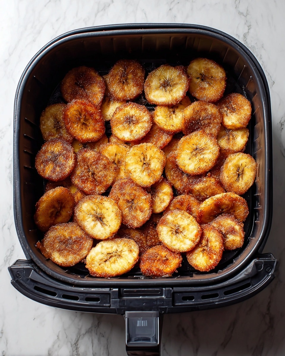 The image shows a black air fryer basket filled with one layer of round, golden-brown fried plantain slices. Each slice has a slightly crispy, caramelized texture with a light sprinkle of cinnamon and sugar, giving a shiny and slightly grainy look on top. The air fryer basket contrasts with the white marbled surface below it. photo taken with an iphone --ar 4:5 --v 7