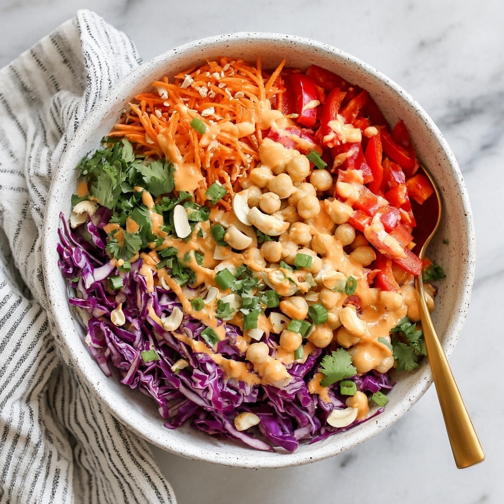 The image shows a white speckled bowl filled with a colorful salad that has several layers. The bottom layer consists of chopped purple cabbage with a crinkly texture, mixed with bright orange shredded carrots. Above this is a layer of chickpeas, tender and round, scattered throughout. There are chunks of red bell pepper adding bold red color and a mix of chopped green onions and fresh green cilantro leaves scattered on top. Some cashew nuts add a light beige and slightly shiny texture. The salad is drizzled with a creamy orange dressing that partially coats the ingredients, and a gold spoon is partially submerged in the bowl. The bowl rests on a white marbled surface next to a striped cloth. Photo taken with an iphone --ar 4:5 --v 7