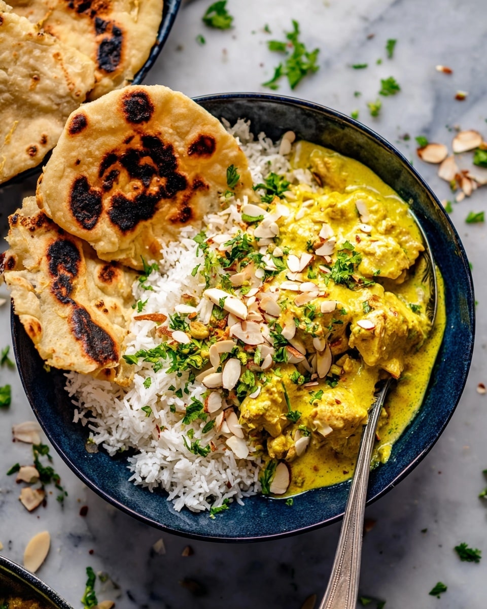 A bowl shows a dish with three parts: on the left, two pieces of round, brownish flatbread with dark char marks and a soft texture resting on the side; in the middle and right side, there is a pile of white rice with individual grains visible and topped with slivered toasted almonds and green chopped herbs; covering part of the rice and flatbread is a thick yellow curry sauce with soft, chunky pieces of meat or tofu, sprinkled with more green herbs and almonds. The bowl is dark blue, set on a white marbled surface with some scattered herbs and almond slices visible. A silver fork is placed on the right side inside the bowl. Photo taken with an iphone --ar 4:5 --v 7